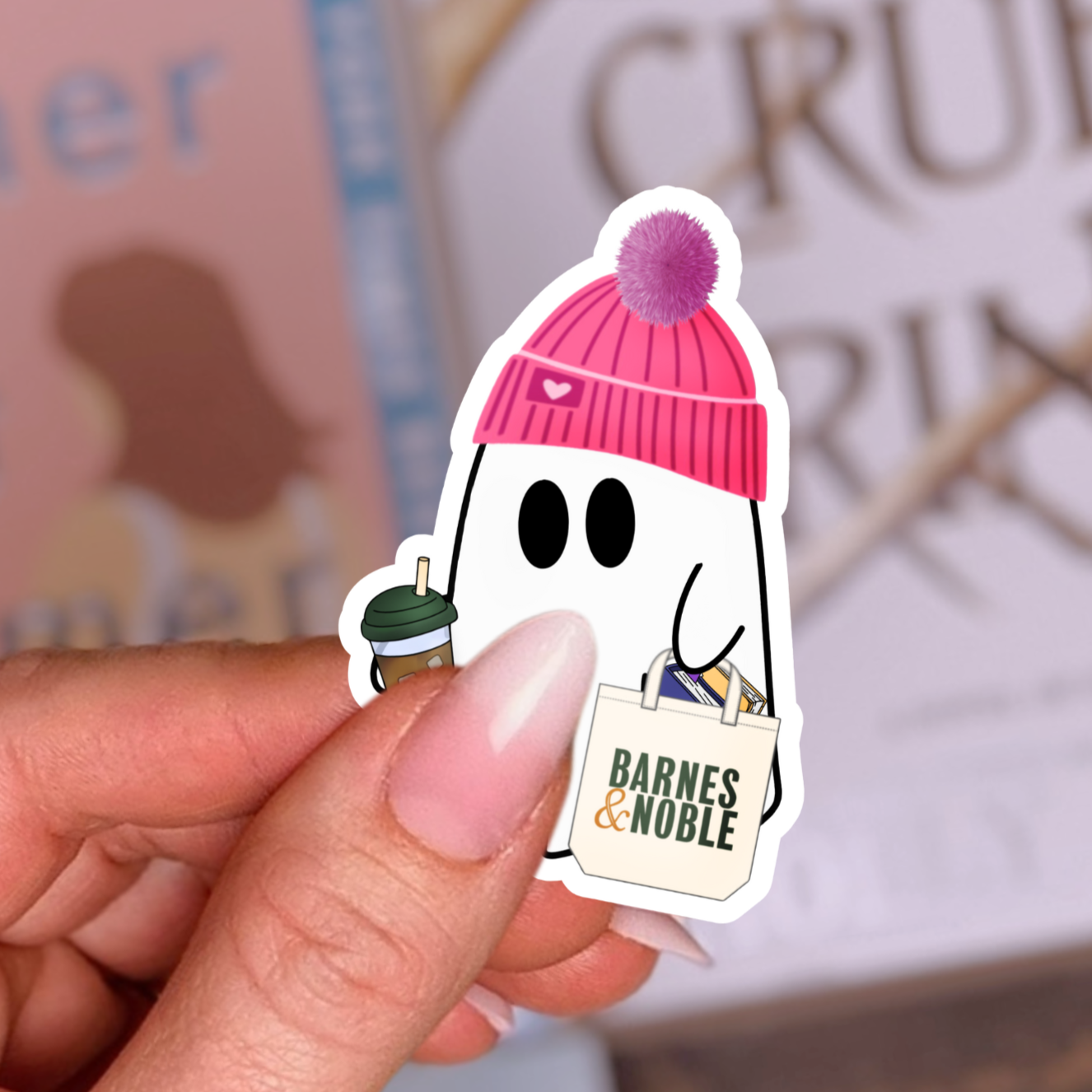 Hand holding a sticker of a ghost in a pink beanie with a Barnes & Noble book, books in the background